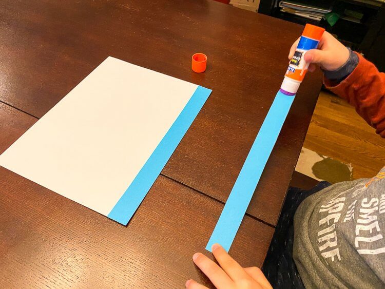 Child gluing blue strips of paper onto a white sheet of paper to make a Greek Flag.