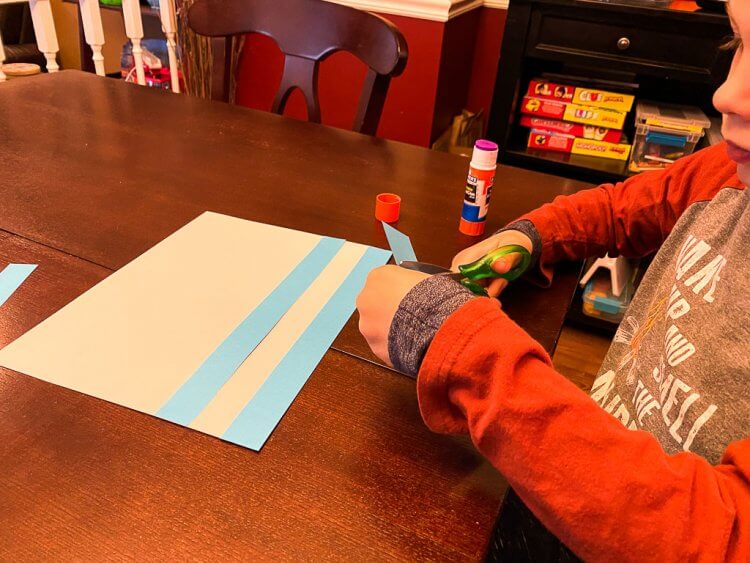 Child with orange sleeves gluing blue strips of paper to a white sheet of paper to make a Greek flag.