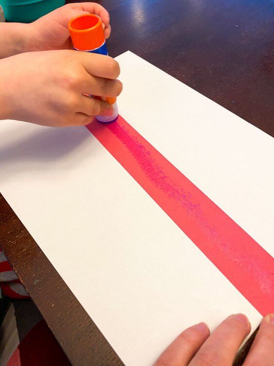 A child's hand gluing a red strip of paper to a white sheet of paper.