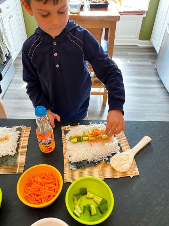Little boy adding carrots, avocado and cucumbers onto his sushi roll.