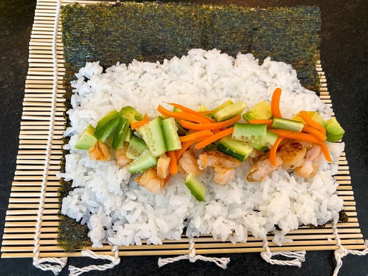 Sushi with white rice and a mix of vegetables laying on a seaweed sheet over a bamboo mat.