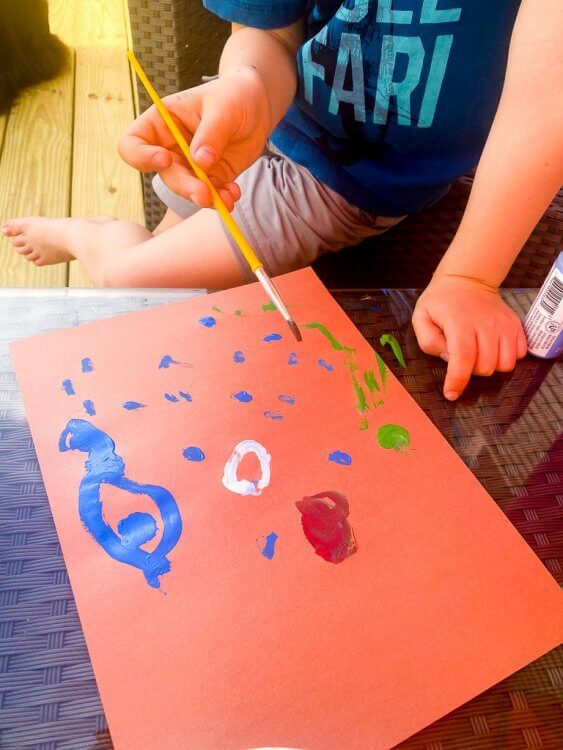 Little boy painting blue, green and white dots on orange paper to make a Japanese fish kite.