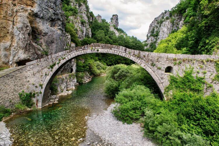 Photos of a bridge in modern day Greece. The bridge is over a river with lush green and high mountains.